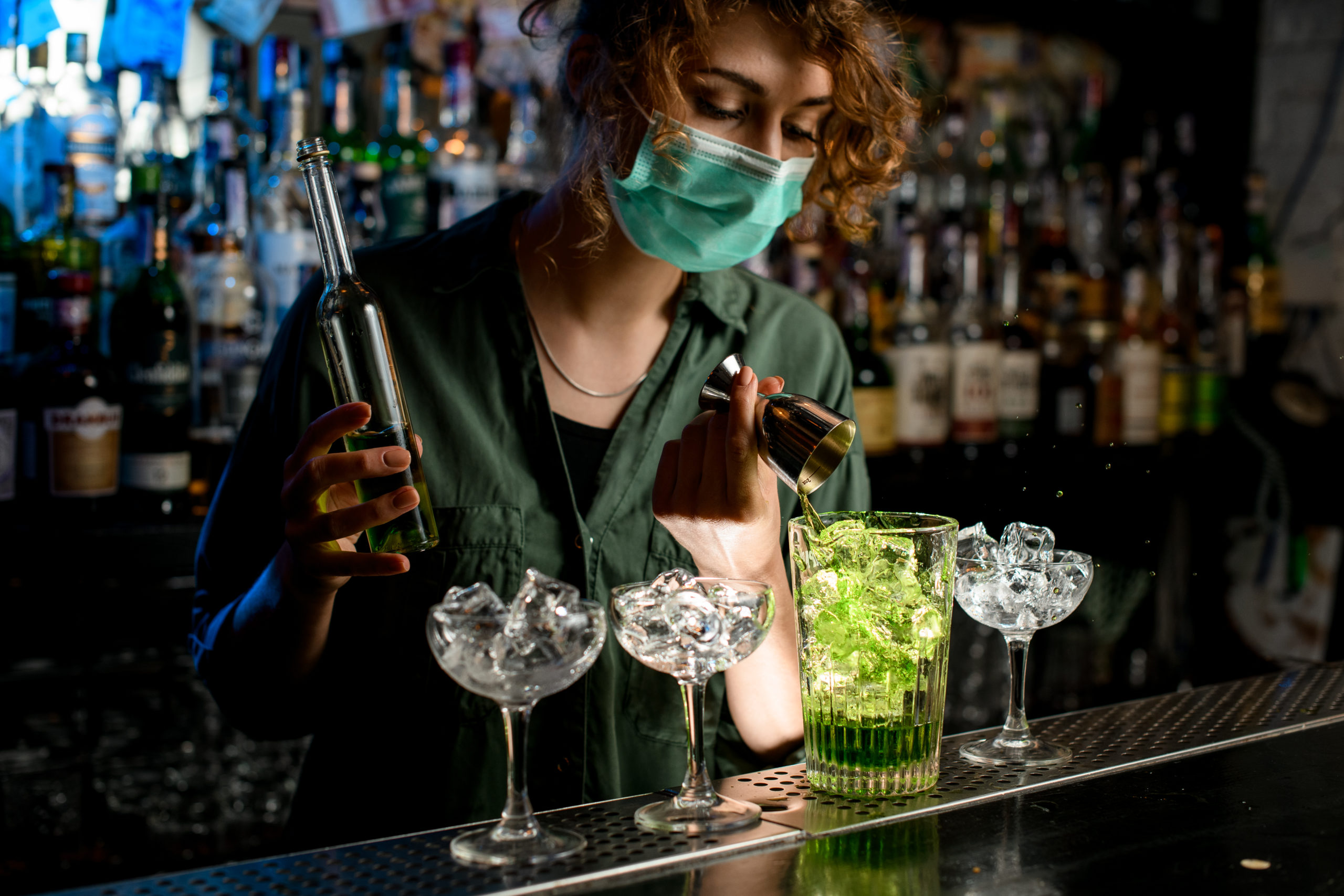 Young bartender girl in a medical mask pour green liquid from beaker into glass with ice.