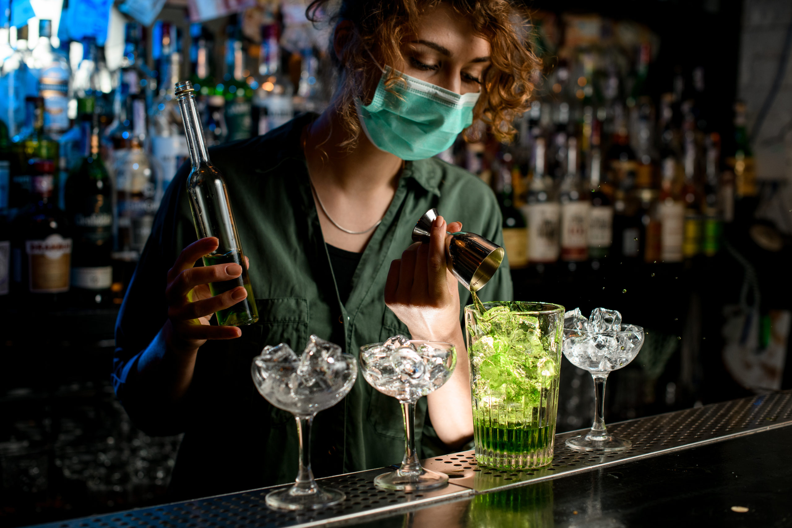 Young bartender girl in a medical mask pour green liquid from beaker into glass with ice. Young bartender girl in a medical mask pour green liquid from beaker into glass with ice.